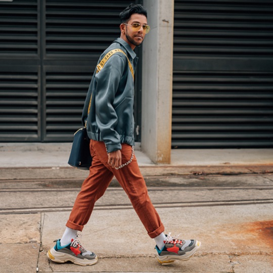 Thinesh poses for camera in his street wear (sneakers, wallet chain, rolled chinos, cross body bag with yellow strap, yellow lensed glasses) striding across the pavement in an urban looking environment, Thinesh is a queer man of colour from Eelam Tamil background